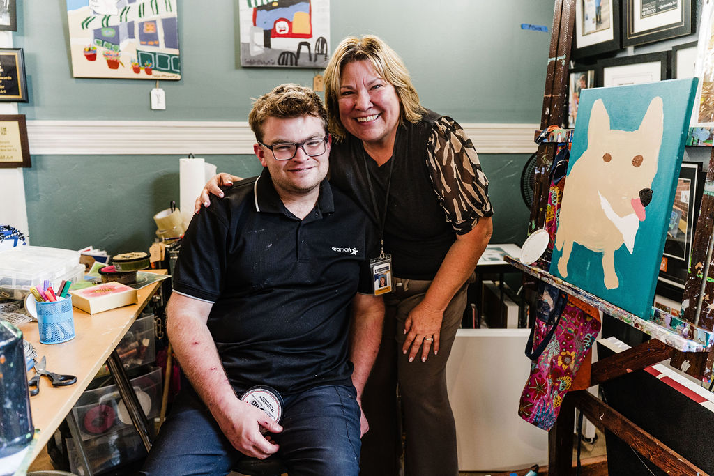 A color photograph of a man and a woman close together in an art studio filled with colorful paintings of pets