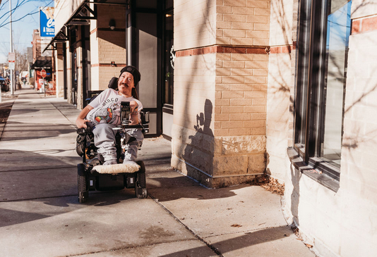 A color photograph of a woman with her back to the camera wearing a bright pink shirt that reads it's a great day for inclusion