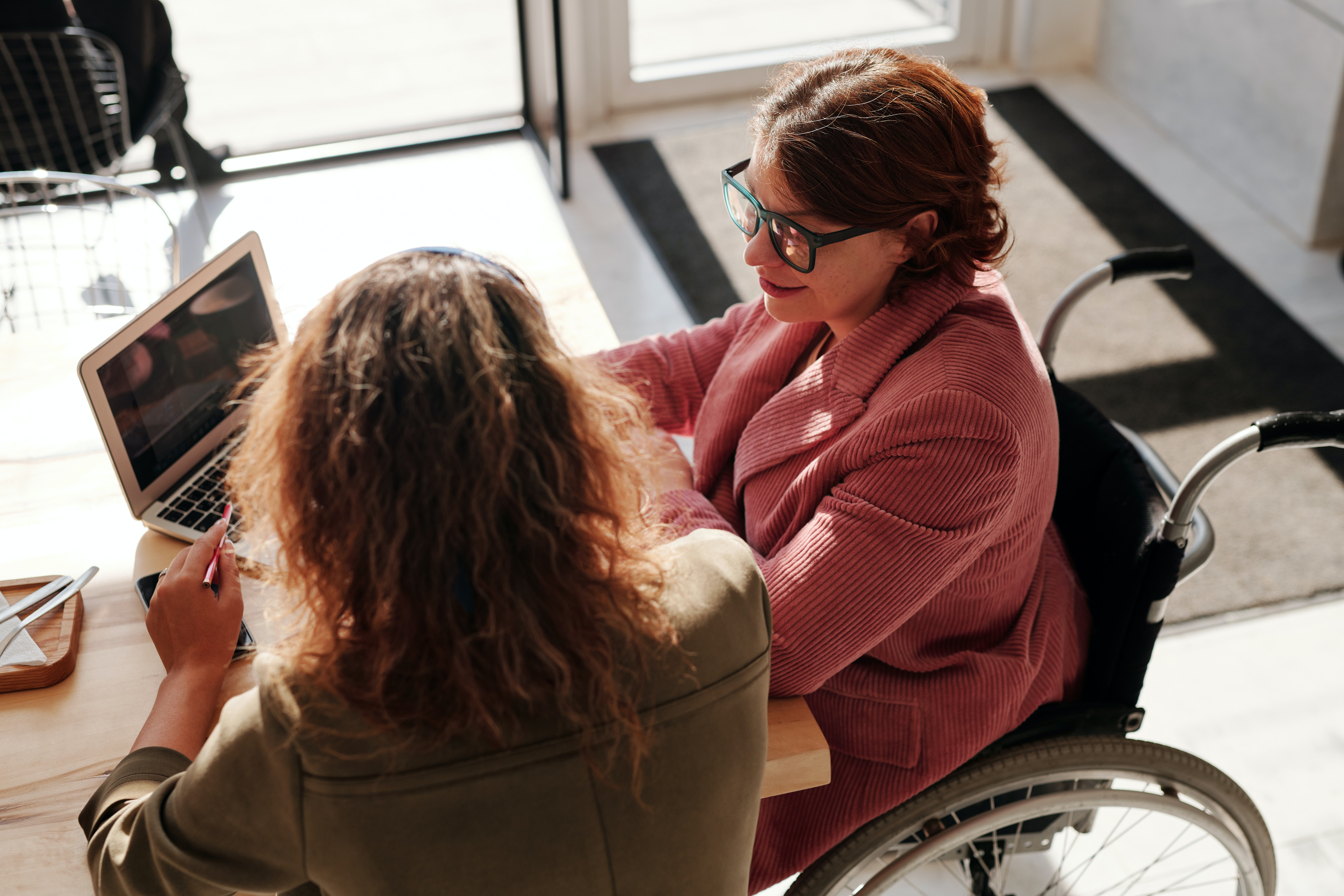 Two women sitting at a table looking over a laptop. One woman is sitting in a wheelchair and the other is sitting in a chair.