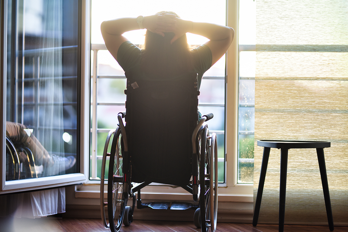 Photo of a woman in a wheelchair, in front of a window, with her back to the camera.